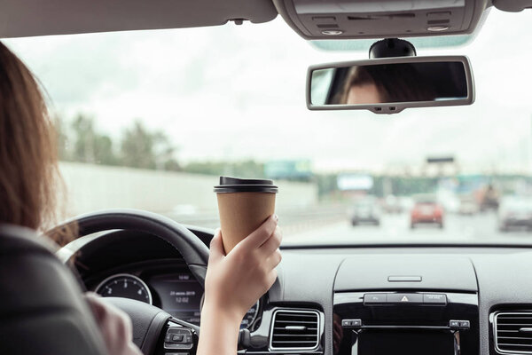 Woman is driving on the autobahn and holding paper cup of coffee at the morning . View from the back seat of the car 