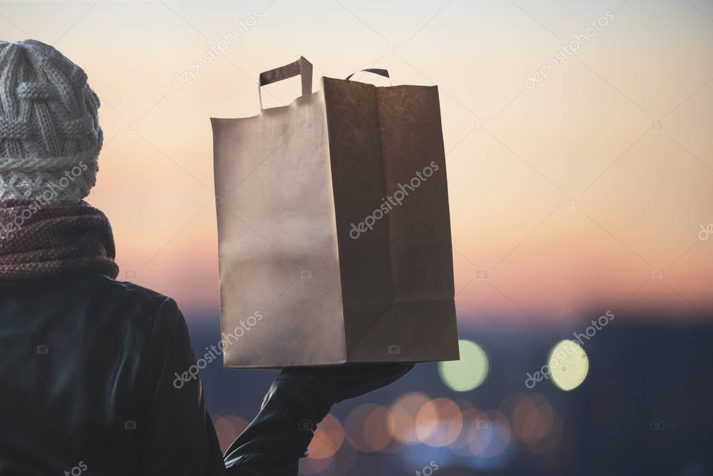 A woman in a winter clothes with a paper bag on the background of the evening city. 24 hours delivery. Concept