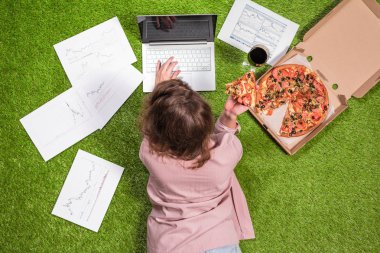 Gazeteye basılmış grafik ve grafiklerle dizüstü bilgisayar kullanarak çimüzerinde çalışan pizza ve bir kadeh şarapla öğle yemeği alan kadın.