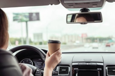 Kadın sabah otoyolda araba sürüyor ve elinde bir fincan kağıt kahve tutuyor. Arabanın arka koltuğundan bak. 
