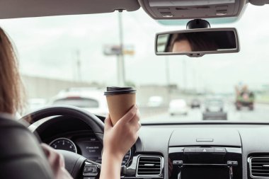 Kadın sabah otoyolda araba sürüyor ve elinde bir fincan kağıt kahve tutuyor. Arabanın arka koltuğundan bak. 