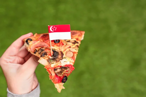 つまようじの形でシンガポールの旗を持つ女性の手のピザのスライス 緑の草の上で昼食 — ストック写真