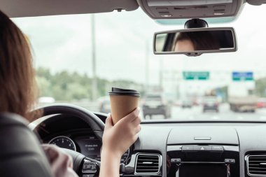 Kadın otobanda sürüyor ve sabahları kağıt bir fincan kahve tutuyor. Arabanın arka koltuğundan görünüm 