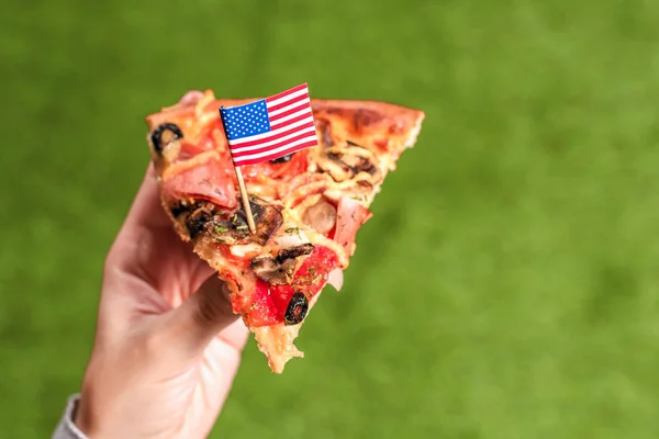 つまようじの形でアメリカの旗を持つ女性の手のピザのスライス 緑の草の上で昼食 — ストック写真