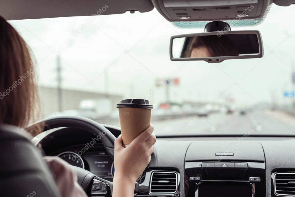 woman is driving on the highway and holding paper cup of coffee at the morning . View from the back seat of the car 