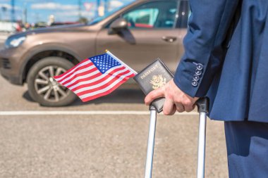Mavi takım elbiseli bir adam bavul bayrağı ve Amerikan pasaportu ile havaalanı otoparkında, arabasının arka planında. İş gezisi konsepti