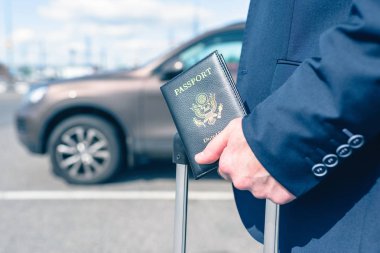 Mavi takım elbiseli, valizli ve Amerikan pasaportlu bir adam havaalanında arabasının arka planına park etmiş. İş gezisi konsepti