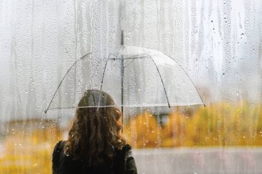 Bir kadın silueti, şeffaf bir şemsiyeyle ıslak pencereden yağmur damlalarıyla. Sonbahar
