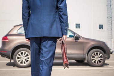 Orta yaşlı, gri saçlı, mavi takım elbiseli, kahverengi çantalı bir işadamı özel park yerinde arabasına doğru yürüyor.