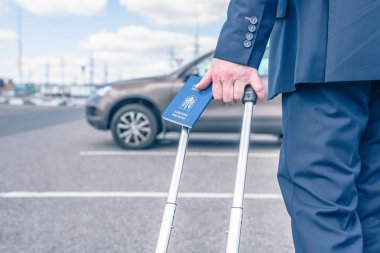 Mavi takım elbiseli, valizli ve Ukrayna pasaportlu bir adam havaalanı otoparkında, arabasının arka planında. İş gezisi konsepti