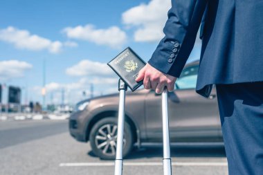 Mavi takım elbiseli, valizli ve Amerikan pasaportlu bir adam havaalanında arabasının arka planına park etmiş. İş gezisi konsepti