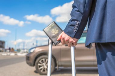 Mavi takım elbiseli, valizli ve Amerikan pasaportlu bir adam havaalanında arabasının arka planına park etmiş. İş gezisi konsepti