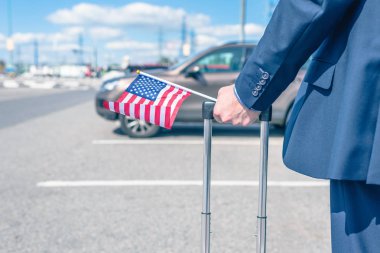 Mavi takım elbiseli, bavullu ve Amerikan bayrağı taşıyan bir adam havaalanında, arabasının arka planında. konsept