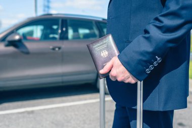 Mavi takım elbiseli, valizli ve Almanya pasaportlu bir adam havaalanındaki park alanında, arabasının arka planında. İş gezisi konsepti