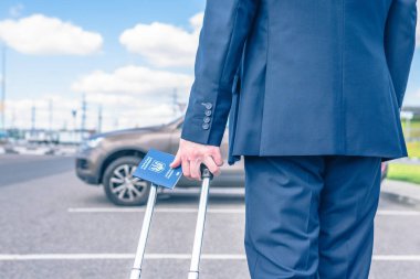 Mavi takım elbiseli, valizli ve Ukrayna pasaportlu bir adam havaalanı otoparkında, arabasının arka planında. İş gezisi konsepti