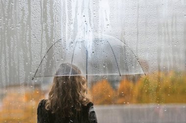 Bir kadın silueti, şeffaf bir şemsiyeyle ıslak pencereden yağmur damlalarıyla. Sonbahar