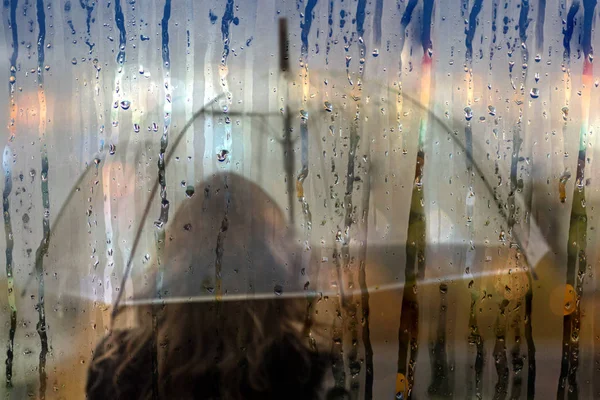 A woman silhouette with transparent umbrella through wet window with drops of rain in night town with night lights. Autumn