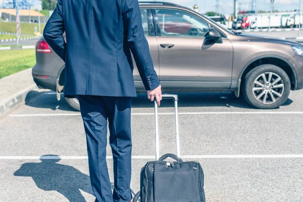 Man in a blue suit with a suitcase at the airport parking on the backdrop of his car. Business trip concept