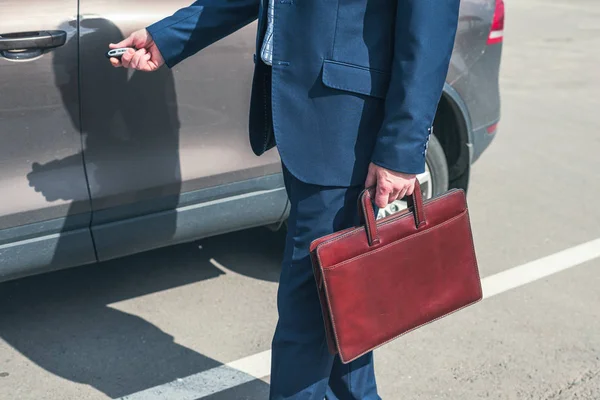 Gray-haired middle aged businessman in a blue suit and briefcase pushing button on remote control car key