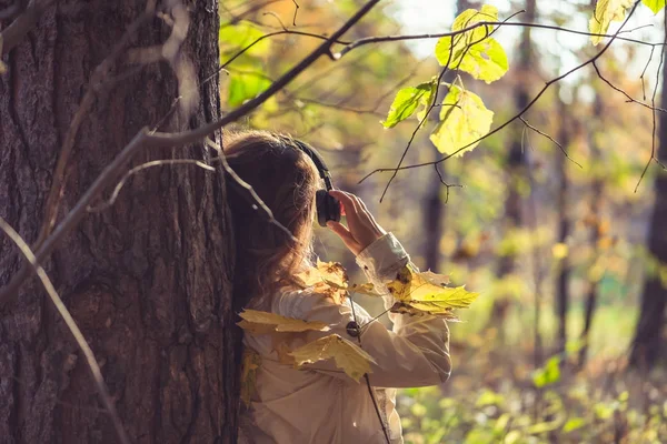 Kulaklıklı bir kadın parkta büyük bir akçaağacın yanında duruyor ve sonbahar ormanında müzik dinliyor. Kavram. Hint yaz sezonu