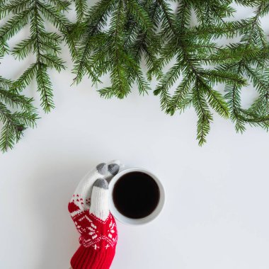 Beyaz bir masanın üzerinde ladin dallarında bir fincan sade kahve ile kış eldivenleri giymiş kadın eller. Noel ve Yeni Yıl konsepti. Kahve sanatı. Düz yerde, üst tarafta. Instagram
