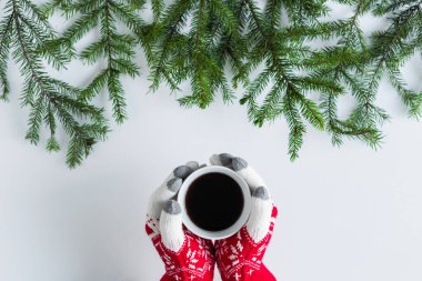Beyaz bir masanın üzerinde ladin dallarında bir fincan sade kahve ile kış eldivenleri giymiş kadın eller. Noel ve Yeni Yıl konsepti. Kahve sanatı. Düz yatış, üst görünüm. 