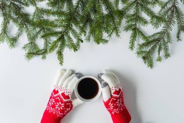 Beyaz bir masanın üzerinde ladin dallarında bir fincan sade kahve ile kış eldivenleri giymiş kadın eller. Noel ve Yeni Yıl konsepti. Kahve sanatı. Düz yatış, üst görünüm. 