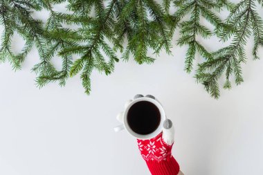 Beyaz bir masanın üzerinde ladin dallarında bir fincan sade kahve ile kış eldivenleri giymiş kadın eller. Noel ve Yeni Yıl konsepti. Kahve sanatı. Düz yatış, üst görünüm. 