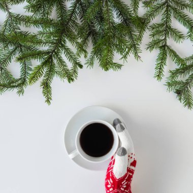 Beyaz bir masanın üzerinde ladin dallarında bir fincan sade kahve ile kış eldivenleri giymiş kadın eller. Noel ve Yeni Yıl konsepti. Kahve sanatı. Düz yerde, üst tarafta. Instagram