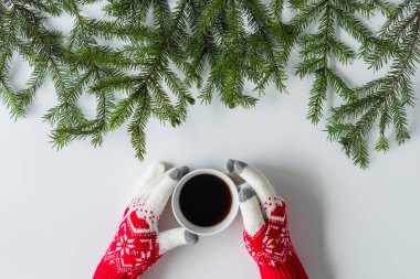 Beyaz bir masanın üzerinde ladin dallarında bir fincan sade kahve ile kış eldivenleri giymiş kadın eller. Noel ve Yeni Yıl konsepti. Kahve sanatı. Düz yatış, üst görünüm. 