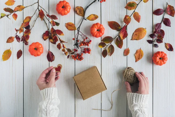 Woman packing gift in a box of kraft paper on white retro wood boards. leaves, rowan and small pumpkins. Thanksgiving. Autumn, fall concept. Flat lay, top view.