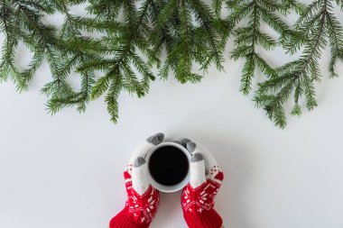 Beyaz bir masanın üzerinde ladin dallarında bir fincan sade kahve ile kış eldivenleri giymiş kadın eller. Noel ve Yeni Yıl konsepti. Kahve sanatı. Düz yatış, üst görünüm. 