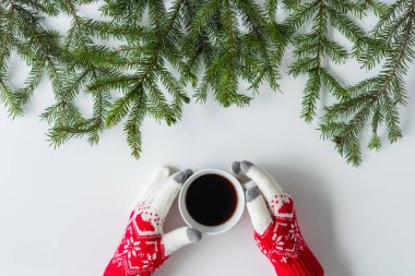 Beyaz bir masanın üzerinde ladin dallarında bir fincan sade kahve ile kış eldivenleri giymiş kadın eller. Noel ve Yeni Yıl konsepti. Kahve sanatı. Düz yatış, üst görünüm. 