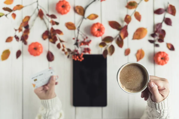 Woman with tablet and credit (discount) card cup on white retro wood boards. Coffee, tablet, leaves, rowan and small pumpkins . Autumn, fall concept. Flat lay, top view.