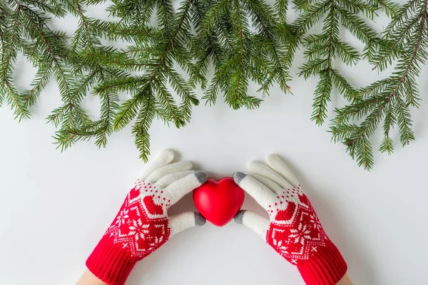 Female hands in the winter gloves with big red heart in spruce branches on a white table. Christmas and New Year concept.  Flat lay, top view.