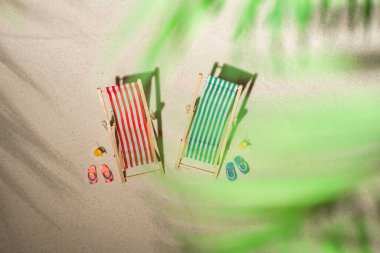 Hava manzaralı iki iskemle, bir bardak meyve suyu, palmiye dalları ve gölge palmiye dalları arasında kum üzerinde parmak arası terlikler. Seyahat, tatil, yaz tatili, romantik konsept. Üst görünüm.