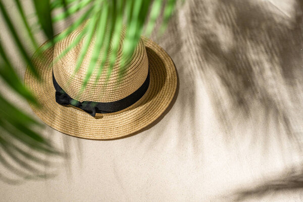 Summer straw hat on sand through the branches of palm and shadow palm tree. Travel, holiday, summertime concept. Top view.