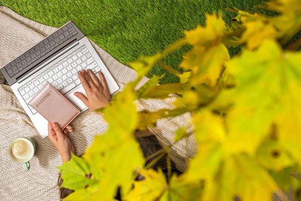 Autumn. Woman on the plaid with laptop and passport, cup of coffee under tree, branch of maple leaves on the green grass in the park. Travel, booking concept. Top view