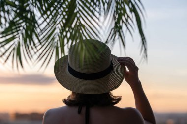 Hasır şapkalı güzel bir kızın, palmiye dallarında denizin arka planına bakan görüntüsü. Gün batımı kumsalı. Yaz ve özgürlük kavramı.