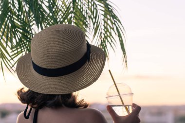 Palmiye dallarında denizin arka planında, hasır bir şapkayla soğuk su içen güzel bir kızın arka plan görüntüsü. Gün batımı kumsalı. Yaz ve özgürlük kavramı.
