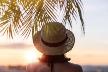 Hasır şapkalı, yalnız, üzgün bir kadının, ufukta batan güneşin karşısında dikilirkenki arka görüntüsü..