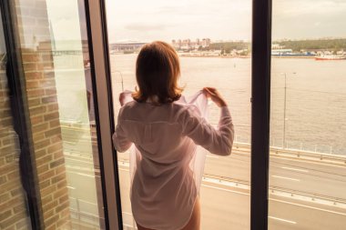 Şeffaf geceliğiyle, gökdelenlerin açık panoramik penceresinin yanında duran ve eğlenen genç ve güzel bir kadının arka görüntüsü. Kasaba ve yol boyunca kadın kolları.