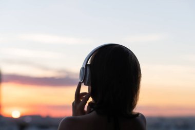 Arkaya bakan, kulaklık takan, gün batımında sahilde müzik dinleyen rahat bir kadın silueti.