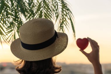 Hasır şapkalı güzel bir kızın arka planda, palmiye dallarında denizin arka planına karşı kırmızı kalbi tutuşu. Gün batımı kumsalı. Yaz ve özgürlük kavramı.