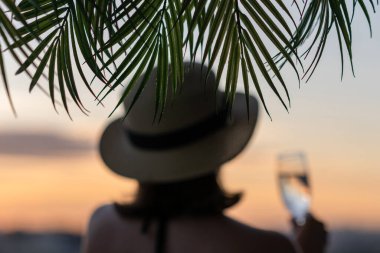 Yalnız, bulanık, bulanık, bir bardak şampanyası olan, hasır şapkalı, palmiye dallarında denizin arka planına bakan bir kız..