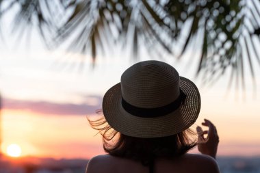 Hasır şapkalı güzel bir kızın, palmiye dallarında denizin arka planına bakan görüntüsü. Gün batımı kumsalı. Yaz ve özgürlük kavramı.