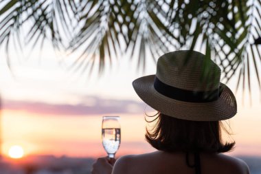 Yalnız ve güzel bir kızın arka planında hasır bir şapkayla palmiye dallarında denizin arka planına bakan bir kız. Gün batımı kumsalı. Yaz ve özgürlük kavramı.