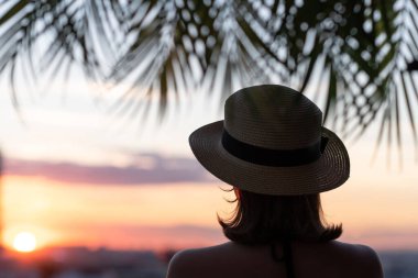 Hasır şapkalı güzel bir kızın, palmiye dallarında denizin arka planına bakan görüntüsü. Gün batımı kumsalı. Yaz ve özgürlük kavramı.