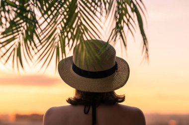 Hasır şapkalı güzel bir kızın, palmiye dallarında denizin arka planına bakan görüntüsü. Gün batımı kumsalı. Yaz ve özgürlük kavramı.