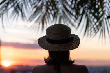 Hasır şapkalı güzel bir kızın, palmiye dallarında denizin arka planına bakan görüntüsü. Gün batımı kumsalı. Yaz ve özgürlük kavramı.